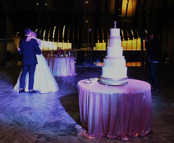 Jeremy and Stacie Hurtt dance in the distance behind their wedding cake to a songs spun by DJSean Hearn at their reception at Crystal Bridges Museum in Bentonville Northwest Arkansas.